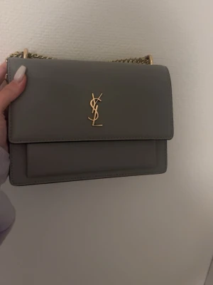 Grå axelväska från Saint Laurent - Säljer en stilren grå axelväska från Saint Laurent med guldfärgad kedja och ikoniskt YSL-emblem framtill. Väskan är i slätt skinn och har magnetlås samt en praktisk ficka på baksidan. Perfekt storlek för det viktigaste och ger en lyxig touch till din outfit.
