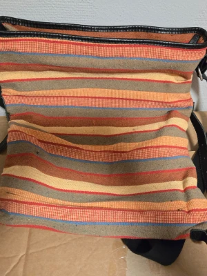 Randig axelväska  - Säljer en färgglad axelväska med randigt mönster i orange, beige, rött, blått och brunt. Väskan har svart kantband, justerbar axelrem och flera fack med dragkedja inuti.