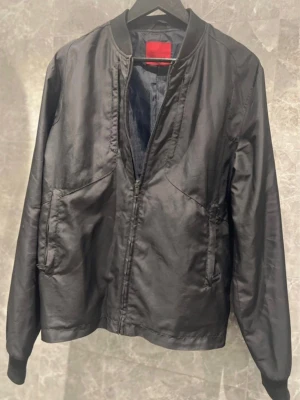 Svart bomberjacka med dragkedja - Säljer en svart bomberjacka i polyester med klassisk ribbad krage, muddar och nederkant. Jackan har dragkedja framtill och två snedställda fickor. Enkel och stilren design som passar till det mesta. Perfekt för dig som gillar en clean look.