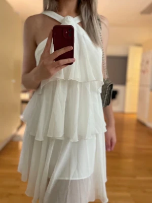 Vit omlottklänning med volanger - Säljer en superfin vit klänning med omlott och lager på lager-volanger. Klänningen har ett snyggt halterneckband och är ärmlös, vilket ger en luftig och elegant känsla. Perfekt för sommarens alla tillfällen!