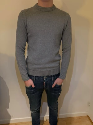 Grå stickad tröja från Dobber - En stilren grå stickad tröja från Dobber i storlek S. Tröjan har rund halsringning, ribbade muddar och är långärmad. Perfekt för lager-på-lager och funkar till både jeans och chinos. Enkel och klassisk design som är lätt att matcha.