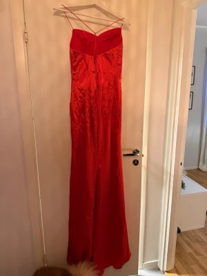 Röd långklänning med snörning - Snygg röd långklänning i glansigt material med tunna axelband och snörning i ryggen. Klänningen har en draperad framsida och figurnära passform som ger en elegant siluett. Perfekt för speciella tillfällen när du vill sticka ut. Var tänk att ha till balen men hade en annan så helt oanvänd 