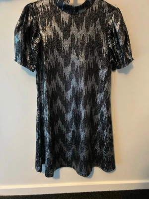 Glittrig svart och silver kortklänning - Säljer en glittrig kortklänning i svart och silver med paljetter och geometriskt mönster. Klänningen har puffiga korta ärmar, hög krage och knäppning i nacken. Perfekt för dig som vill sticka ut med en riktigt snygg look.