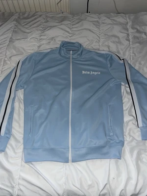 Ljusblå Palm Angels track jacket - Säljer en ljusblå track jacket från Palm Angels med vita och svarta ränder längs ärmarna. Jackan har hög krage, hel dragkedja framtill och två fickor. Materialet är polyester och jackan har en clean, sportig vibe med logga på bröstet.