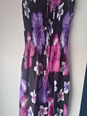 Blommig axelbandslös klänning - Säljer en axelbandslös klänning med smock upptill och stort blommönster i lila, rosa och vitt på svart botten. Klänningen har en avslappnad passform och knytband framtill. Köpt på en marknad i Spanien för några år sedan så vet ej storlek men skulle gissa på xs eller xxs