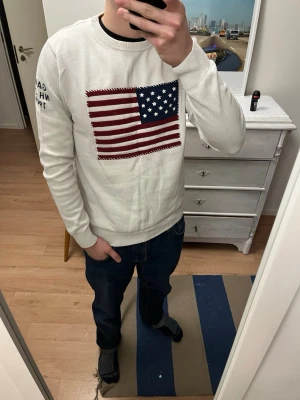 Vit stickad tröja från GANT med USA-flagga - Vit stickad tröja från GANT med stor USA-flagga broderad på bröstet och tryck på ena ärmen. Rund hals och ribbade muddar vid ärmslut och nederkant. Perfekt för dig som gillar klassisk preppy-stil och vill sticka ut med en snygg detalj.
