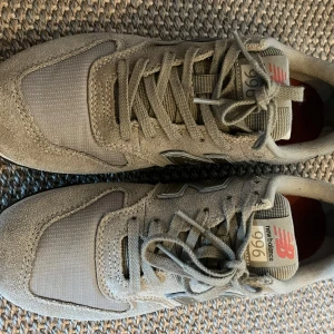 Beige New Balance 996 sneakers - Säljer ett par beige New Balance 996 sneakers i storlek 37.5. Skorna har en snygg mix av mocka och textil, med klassisk N-logga på sidan och röda detaljer vid hälen. De har snörning och en platt sula för skön vardagsstil.