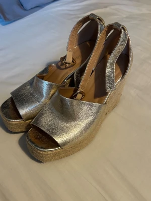 Guldiga platåsandaler med rem - Snygga sandaler i metallic guld med bred rem över foten och öppen tå. Skorna har en hög platåsula i flätad jute och justerbar ankelrem med spänne. Perfekta för sommarens alla tillfällen när du vill sticka ut lite extra.