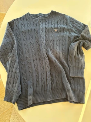 Mörkgrå kabelstickad tröja från Gant - Säljer en mörkgrå kabelstickad tröja från Gant i premium bomull. Tröjan har rund halsringning, ribbade muddar och Gant-logga broderad på bröstet. Perfekt för dig som gillar klassisk och stilren look.