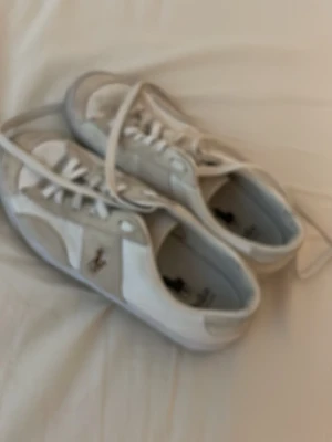 Vita sneakers från Polo Ralph Lauren - Säljer ett par klassiska vita sneakers från Polo Ralph Lauren med den ikoniska hästloggan i brunt på sidan. Skorna har snörning, rund tå och är tillverkade i en mix av skinn och syntet. Perfekta till jeans eller shorts för en clean och stilren look.Är villig att dra ner priset