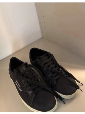 Svarta Saint Laurent sneakers - Svarta sneakers från Saint Laurent med vit logotyp på sidan. Skorna har snörning, rund tå och vit platt sula. Materialet är canvas och skinn, vilket ger en cool och stilren vibe. Levereras med originalkartong, dustbag och extra skosnören. Skicket är 8/10 de är bara naturligt slitag men inget som påverkar utseendet, köpta på storeenda, priset är ej hugget i sten