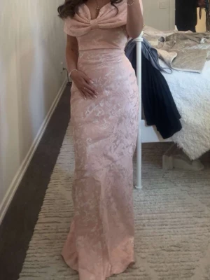 Ljusrosa långklänning med snörning - Supersnygg ljusrosa långklänning med offshoulder och stor rosett framtill. Klänningen har ett subtilt blommigt mönster och snörning i ryggen för extra snygg passform. Perfekt för dig som vill sticka ut med en elegant och feminin look.