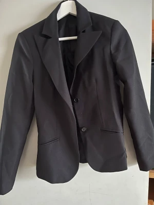 Svart kavaj med knappar - Snygg svart kavaj med klassisk krage och två knappar framtill. Figursydd modell med långa ärmar och två fickor. Perfekt för att lyfta vilken outfit som helst.