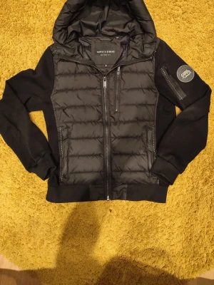 cardigan jacka Supply & Demand - Snygg svart cardiganjacka från Supply & Demand med huva och dragkedja framtill. Jackan har quiltade partier på bröst och rygg, samt släta ärmar och muddar. Märkespatch på ena ärmen och flera praktiska fickor med dragkedja.