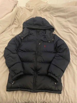 Mörkblå pufferjacka från Polo Ralph Lauren - Säljer en mörkblå pufferjacka från Polo Ralph Lauren i storlek XL. Jackan har huva, dragkedja framtill och en röd broderad logga på bröstet. Fylld med dunliknande material och har praktiska fickor med dragkedja. Perfekt för kalla dagar.