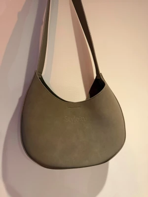 Beige axelväska  - Säljer en stilren beige axelväska från Amazing Seven som vi har fått in från Stylein! Väskan heter mini yardly bag i färgen Brown Matte. Perfekt för dig som gillar enkel och clean design! Är själv anställd på Amazing Seven och har köpt via min chef som ett p-köp! Tygpåsen är kvar! Höjd: 23cm, Bredd: 26cm, Axelrem: 50cm. OBS! Gjord av fuskmocka