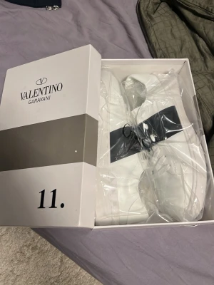 Valentino Garavani vita sneakers strl 45 - Säljer ett par snygga vita Valentino Garavani sneakers med svart band och coola nitar på sidan. Skorna har rund tå, snörning och är tillverkade i skinn. Perfekta för dig som vill ha en lyxig och stilren look med designer vibe.