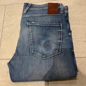 Replay Anbass blå jeans, - Säljer ett par Replay Anbass jeans i klassisk blå tvätt med snygga slitningar och kontrastsömmar. Modellen har fem fickor, normal passform och raka ben. Jeansen är tillverkade i mjukt denim-material med detaljer som röd broderad storlek och Replay-lapp bak. Pris kan diskuteras!