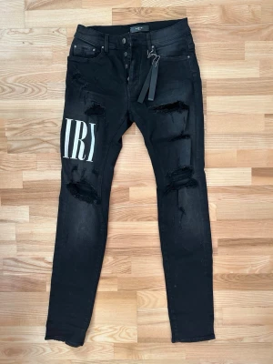Svarta skinny jeans från Amiri storlek 30 - Svarta jeans från Amiri med skinny passform och slitna detaljer på benen. Stora vita tryck med bokstäver på framsidan och baksidan av vänster ben. Klassisk femficksdesign, knappgylf och tagg från Amiri. Materialet är jeans med stretch.
