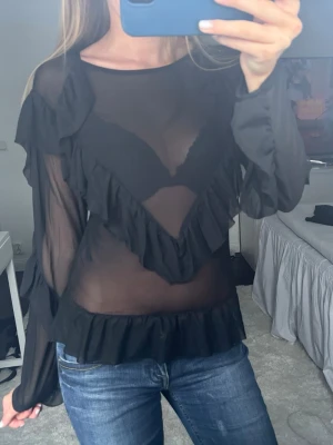 Genomskinlig festtopp  - Säljer en svart genomskinlig blus med långa ärmar och snygga volanger framtill och vid midjan. Storlek M från MSCH Copenhagen.