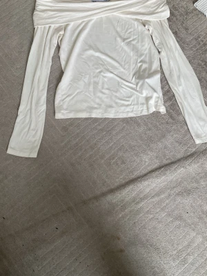 Vit offshoulder långärmad topp - Säljer en vit offshoulder topp med långa ärmar. Toppen har en bred kant upptill som ger en snygg och avslappnad look. Materialet känns mjukt och lätt, perfekt för att styla med jeans eller kjol. Enkel och stilren design som funkar till många outfits.