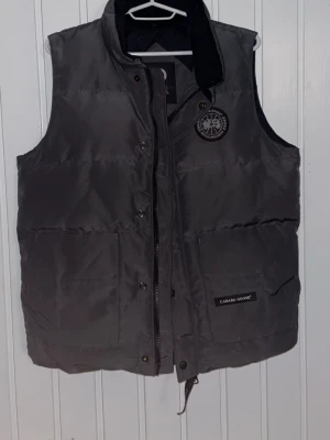 Black label väst från Canada Goose - Säljer en ljusgrå dunväst från Canada Goose med hög krage och två stora fickor framtill. Västen har dragkedja och knappar samt en snygg broderad logga på bröstet. Perfekt för lager-på-lager under kyliga dagar.