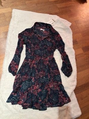 Blommig klänning.  Holly & White - Supersnygg kortklänning från Holly & White med mörkblå botten och blommigt mönster i rött och vitt. Klänningen har långa ärmar, markerad midja och volangkant nertill. Perfekt för dig som gillar en feminin och trendig stil.