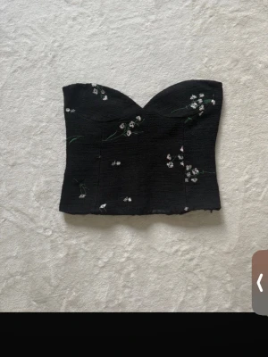 Svart blommig korsett-topp - Säljer en svart korsett-topp med söt blommig brodyr i vitt och ljusrosa. Toppen är axelbandslös och har en hjärtformad överkant som ger en snygg siluett. Perfekt att styla med jeans eller kjol för en trendig look.