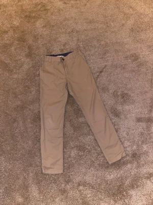 Beige chinos från H&M - Snygga beige chinos från H&M med klassisk passform och knappstängning i midjan. Byxorna har raka ben och är tillverkade i mjuk bomull, vilket gör dem sköna att ha på sig hela dagen. Perfekta för dig som gillar stilrena och enkla plagg.