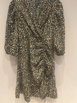 Leopardmönstrad omlottklänning Vero Moda - Snygg kort omlottklänning från Vero Moda med leopardmönster i svart, beige och vitt. Klänningen har v-ringad hals, trekvartsärmar och volangkant nedtill. Materialet är mjuk polyester och modellen har en smickrande rynkning framtill.