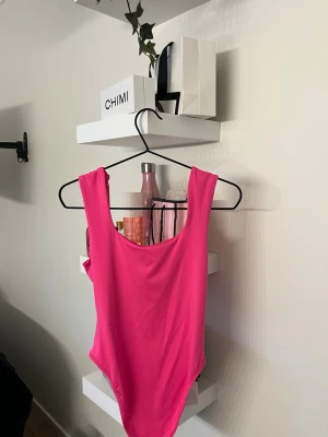 Rosa body  - Säljer denna snygga knallrosa body som är helt oanvänd, den ha breda axelband och djup rundad halsringning. Bodyn har en enkel och stilren design utan mönster eller detaljer, perfekt för dig som gillar färgstarka plagg. Materialet känns mjukt och stretchigt.