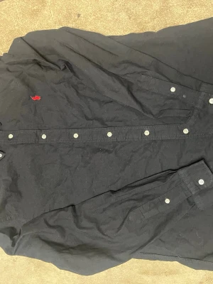 Svart skjorta från Ralph Lauren - Svart skjorta från Ralph Lauren med klassisk krage och vita knappar framtill. På bröstet finns den ikoniska röda Polo-loggan broderad. Skjortan är långärmad och tillverkad i mjuk bomull som känns skön mot huden. Perfekt för dig som gillar stilrena och tidlösa plagg.