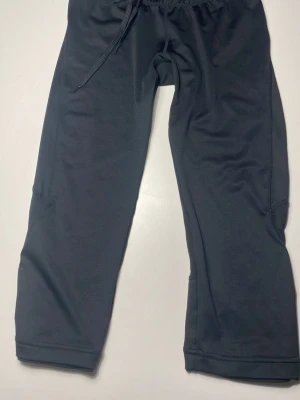 Svarta träningsbyxor med snörning - Svarta träningsbyxor med elastisk midja och snörning, samt dragkedja vid fickan. Byxorna har en loose passform och är tillverkade i mjukt syntetmaterial. Croppad längd ger en modern vibe.