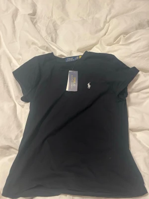 Svart t-shirt från Polo Ralph Lauren - Svart t-shirt från Polo Ralph Lauren med klassisk rund hals och korta ärmar. T-shirten har en vit broderad logga på bröstet och är gjord i mjuk bomull. Perfekt basic med snygg passform och stilren design.