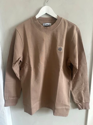 Beige/Brun sweatshirt från Stone Island - Snygg beige sweatshirt från Stone Island med klassisk rund hals och logotyp-patch på bröstet. Tröjan har långa ärmar och ribbade muddar vid ärmslut och nederkant. Tillverkad i mjuk bomull som ger en skön och avslappnad passform.