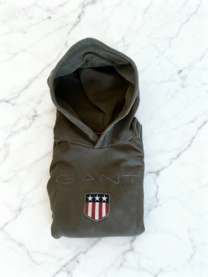 Gant hoodie - Snygg hoodie från GANT i en stilren grön/olivgrön nyans. Tröjan har en tidlös design med diskret GANT-logga på bröstet samt den ikoniska skölden framtill, vilket ger ett exklusivt och sportigt uttryck. Perfekt för både vardag och avslappnade tillfällen. Mjuk och bekväm kvalitet med huva och ribbade muddar för bra passform. Ett mångsidigt plagg som passar året runt och aldrig går ur stil. Skick: mycket bra  Storlek: 158-164 Färg: Grön / olivgrön Märke: GANT