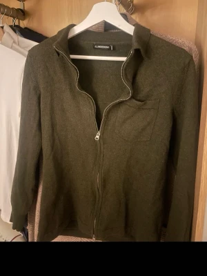 Mörkgrön kofta från J.Lindeberg - Snygg mörkgrön kofta från J.Lindeberg med hel dragkedja framtill och en bröstficka. Koftan har långärmad design och är tillverkad i mjukt material som passar perfekt för kyligare dagar. Stilren och enkel att matcha med olika outfits.