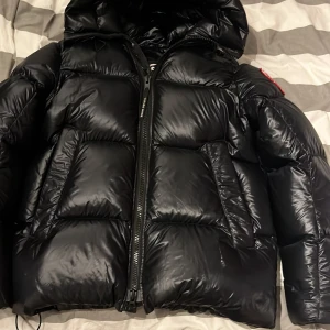 Svart pufferjacka från Canada Goose - Säljer en svart pufferjacka från Canada Goose i storlek M. Jackan har en stor huva, breda quiltade sektioner och dragkedja framtill. Materialet är glansigt och känns riktigt robust, perfekt för kalla dagar. Ikonisk logga på ärmen och snygg passform.