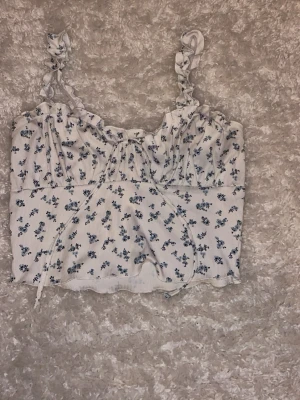 Vit blommig linnetopp med volangband från Shein stl M - Supersöt vit linnetopp med små blå blommor och volangdetaljer på axelbanden i stl M. Toppen har ribbad struktur och en omlottliknande söm framtill. använd fåtal gånger, köparen betalar frakt☺️