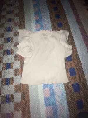 Vit blus med volangärmar - Säljer en söt vit blus med volangdetaljer på ärmarna och rund hals. Blusen har en lätt ribbad struktur och är kortärmad, perfekt för vår och sommar. Passar dig som gillar romantisk och feminin stil.
