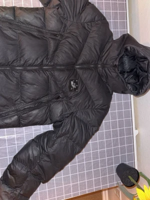 Svart pufferjacka från Nike - Säljer en svart pufferjacka från Nike med huva och två snedställda dragkedjefickor. Jackan har quiltad design och en tydlig Nike-logga på bröstet. Perfekt för kalla dagar och har en modern, sportig vibe. I storleken M