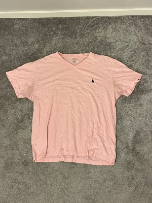 Ljusrosa t-shirt från Polo Ralph Lauren - Säljer en ljusrosa t-shirt från Polo Ralph Lauren med v-ringning och klassisk logga broderad på bröstet. T-shirten är kortärmad och gjord i mjuk bomull, perfekt för en avslappnad och stilren look. Tröjan är använd så slitage finns 