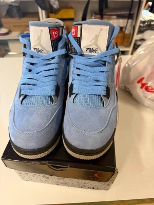 Nike Air Jordan 4 University Blue - Nike Air Jordan 4 University Blue med ljusblå mocka, meshpaneler och svarta samt vita detaljer. Skorna har specklad mellansula, Jumpman-tag och klassisk snörning. Snygg retrostil med grå, svart och vit yttersula. Perfekta för dig som vill sticka ut.