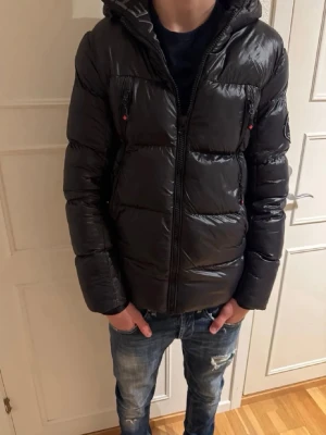 Svart pufferjacka från Zavetti Canada - Snygg svart pufferjacka från Zavetti Canada med glansig finish och broderad logga på ärmen. Jackan har huva, flera dragkedjefickor och en normal passform. Perfekt för kalla dagar och har en modern look som passar streetwear.