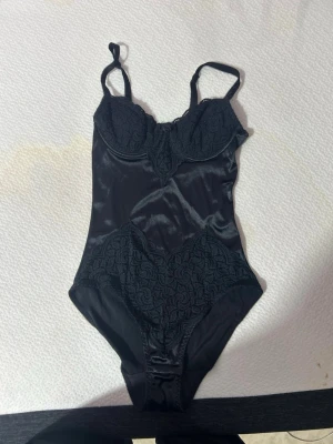Svart korsettbody med spetsdetaljer - Säljer en svart korsettinspirerad body med tunna axelband och snygga spetsdetaljer vid byst och höfter. Materialet är glansigt och mjukt, vilket ger en lyxig känsla. Perfekt för dig som gillar en elegant och stilren look.