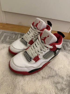 Nike Air Jordan 4 Retro White/Red - Nike Air Jordan 4 Retro sneakers i vit, röd och svart. Skorna har meshpaneler, snörning och ikoniska Jumpman-loggan på tungan. Ovandel i skinn med röda detaljer och svart häl med Nike Air-tryck. Perfekt för dig som vill ha en klassisk och snygg sneaker med streetkänsla.