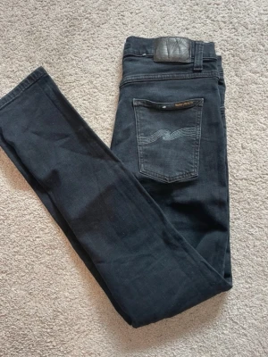 Svarta Nudie Jeans, storlek 29/30 - Säljer ett par svarta jeans från Nudie Jeans i storlek 29/30. Klassisk femficksmodell med snygga ljusblå sömmar på bakfickan och svart läderpatch i midjan. Jeansen har en rak passform och är tillverkade i mjukt bomullsmaterial.