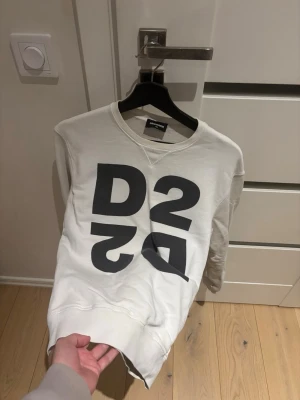 Vit sweatshirt från Dsquared2 - Vit sweatshirt från Dsquared2 med stor svart D2-logga på framsidan. Tröjan har rund hals, långa ärmar och ribbade muddar vid ärmslut och nederkant. Materialet är mjuk bomull som känns skön mot huden. Perfekt för dig som gillar streetwear och tydliga prints.