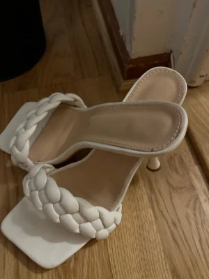 Vita flätade sandaler medt klack - Säljer ett par trendiga vita sandaler med fyrkantig tå och låg klack. Remmen är flätad och ger en snygg detalj. Skorna är tillverkade i skinnimitation och har en öppen design som passar perfekt till varma dagar. Supersnygga till jeans eller klänning.