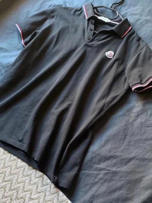 Svart piké från Moncler - Säljer en svart pikétröja från Moncler med klassisk krage och knappar framtill. Pikén har korta ärmar med rödvita ränder på krage och ärmslut samt Moncler-logga på bröstet. Perfekt för dig som gillar stilrena och sportiga plagg. De står L men den sitter som S!! Löser mer bilder om ni vill!!